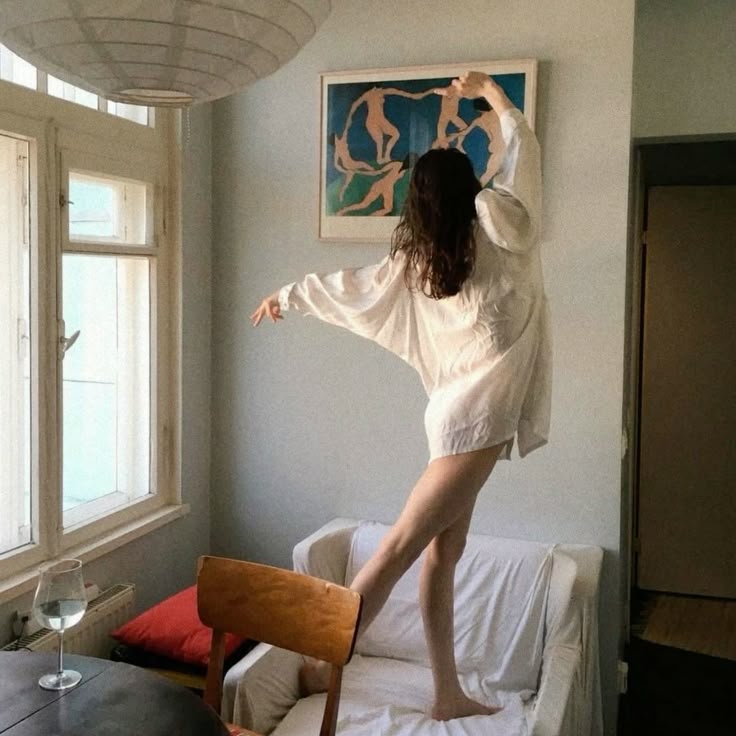 Woman in an oversized white shirt balancing playfully on a couch in a sunlit room with large windows, minimalist decor, and framed artwork.