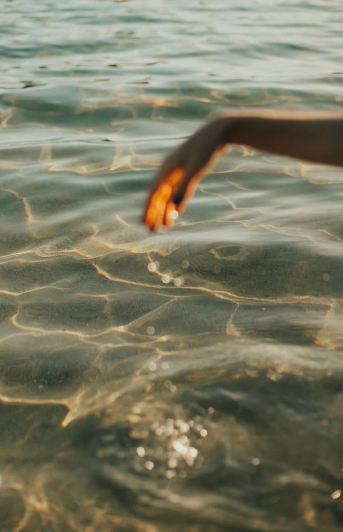 Close-up of a hand gently touching sunlit water, with golden reflections shimmering across the rippling surface.