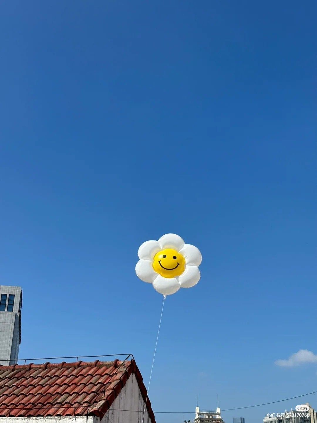 A white daisy-shaped balloon with a yellow smiley face floats against a clear blue sky above red-tiled rooftops and city buildings.