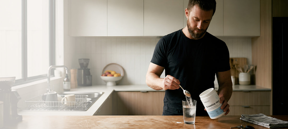 A man in a black t-shirt stands in a modern kitchen, holding a white tub of protein powder Creatine Well and a spoon over a glass of water on a wooden countertop.