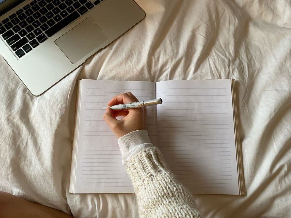 Person writing in a blank notebook beside a laptop, representing tracking food and symptoms in an interstitial cystitis food diary.