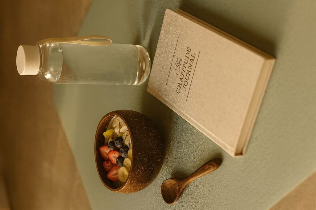 Flat lay of a gratitude journal, smoothie bowl with fruit, wooden spoon, and glass water bottle on a soft surface, representing mindfulness and daily wellness habits.