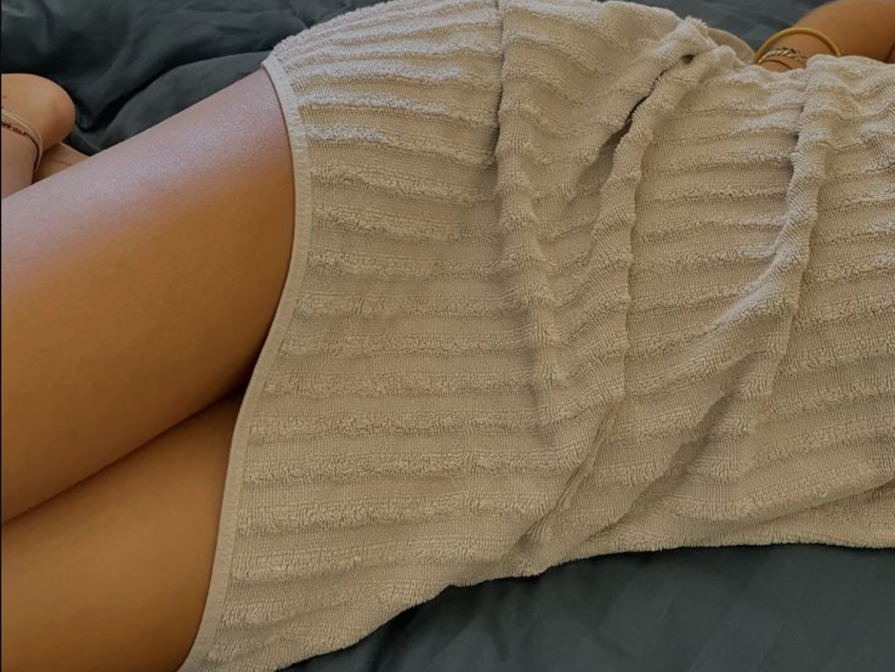 Person lying on a dark bed covered with a beige towel, evoking rest, relaxation, and post-shower calm in a cozy setting.