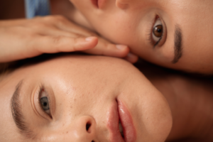 Close-up of two individuals resting side by side, with one person’s face in focus and another gently touching their cheek, highlighting natural skin texture and a calm, intimate moment.