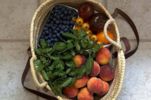 Woven basket filled with fresh produce including blueberries, peaches, tomatoes, herbs, and citrus fruit on a stone surface.