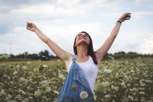 Woman standing in a field of wildflowers with her arms raised, eyes closed, and face turned upward under a cloudy sky.