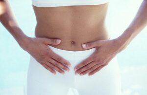 Midsection of a person wearing white clothing, with both hands resting gently on their lower abdomen, suggesting focus on digestion or pelvic health.
