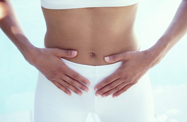 Midsection of a person wearing white clothing, with both hands resting gently on their lower abdomen, suggesting focus on digestion or pelvic health.