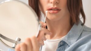 Close-up of a person using a cotton swab to apply product to a cold sore on their lip while looking into a mirror.