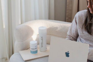 Person seated on a couch with a laptop, next to a tray holding a white Wellex bottle and a blue-and-white supplement bottle.