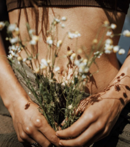 Close-up of a person’s lower abdomen and hands holding small white wildflowers, with warm sunlight casting soft shadows across their skin.