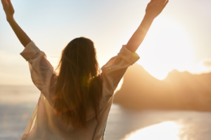 Person standing outdoors with arms raised toward the sky at sunrise or sunset, overlooking water and distant mountains, bathed in warm golden light.