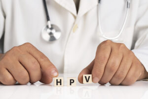 Close-up of a healthcare professional in a white coat with a stethoscope, arranging wooden blocks that spell “HPV” on a table.