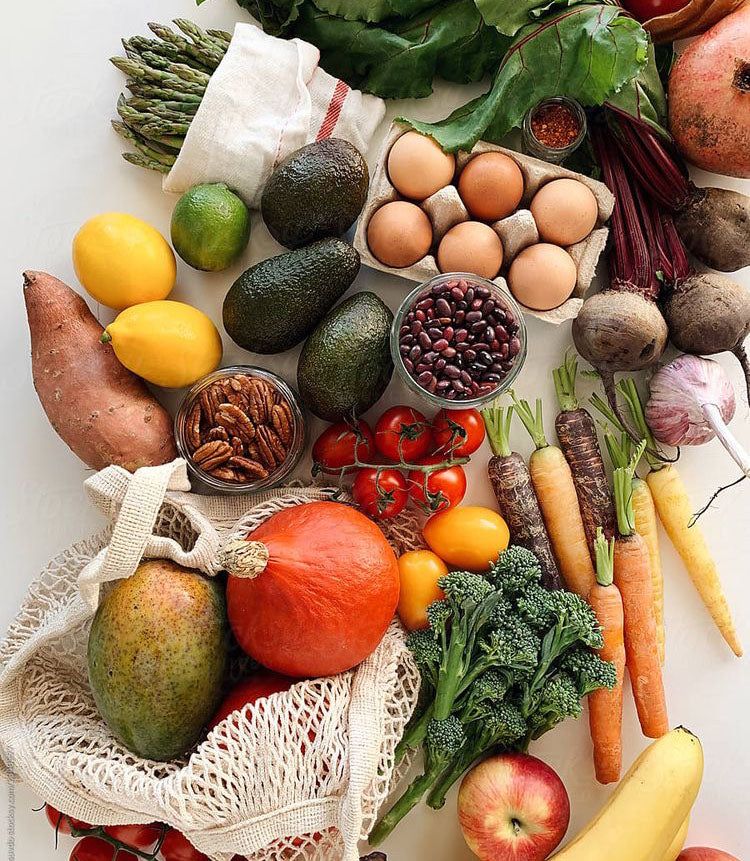 Assortment of fresh whole foods including avocados, eggs, carrots, leafy greens, tomatoes, citrus, broccoli, beans, nuts, and root vegetables arranged on a light surface.