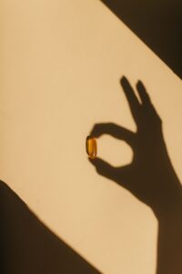 Shadow of a hand holding a golden softgel capsule against warm sunlight and a neutral beige background.