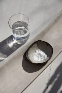 A minimalist wellness scene featuring a small ceramic bowl filled with white creatine powder beside a glass of water on a light stone surface, illuminated by natural sunlight and soft shadows