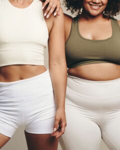 Two women wearing athletic clothing stand closely together, with one placing a hand on the other's shoulder. The image emphasizes body positivity, support, and inclusivity across different body types.