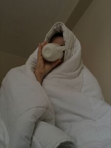 A person wrapped in a white duvet drinks from a white mug, creating a cozy scene that captures the comfort of resting with a warm beverage while feeling under the weather or enjoying a quiet morning indoors.