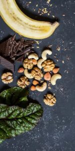 Flat lay of nutrient-rich foods commonly associated with cervical and immune health, including a sliced banana, dark chocolate, walnuts, almonds, cashews, flaxseeds, and leafy greens, arranged on a dark stone background. The image represents a balanced, antioxidant-rich diet that supports overall wellness and immune function.