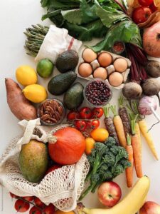 An overhead view of a colorful assortment of whole foods arranged on a white surface, including leafy greens, asparagus, beets, carrots, broccoli, tomatoes, sweet potato, avocado, eggs, beans, pecans, garlic, citrus fruits, banana, apple, pear, mango, and squash, representing a nutrient-rich, balanced diet.