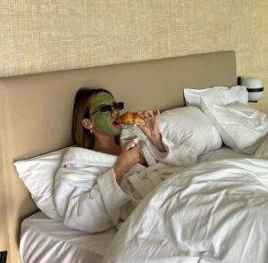 Person relaxing in a hotel bed wearing a white robe, green facial mask, and sunglasses while enjoying a croissant and coffee during a self-care morning.