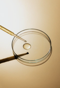 Glass petri dish containing a droplet of clear liquid, with a laboratory pipette positioned above it against a warm beige background.