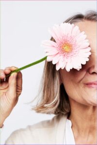 Older woman holding a large pink gerbera daisy over one eye against a light background.