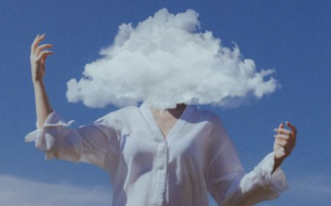 Person in a white shirt standing against a blue sky with a large white cloud obscuring their head, symbolizing brain fog or mental fatigue.