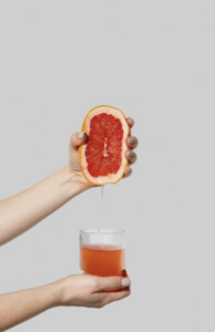Hands squeezing half a grapefruit over a glass of fresh grapefruit juice against a light gray background.