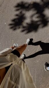 Overhead view of a person walking on a sunlit pavement in athletic wear and white sneakers, with dramatic palm tree shadows cast across the ground.